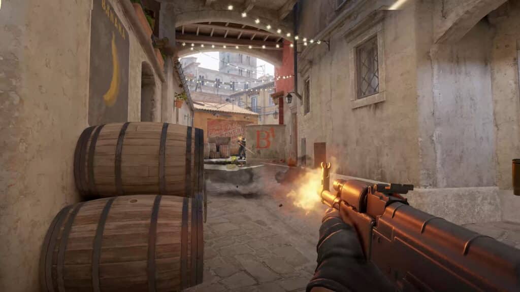 Das Bild zeigt das Gameplay in Counter-Strike 2 auf der Inferno-Karte in einem als „Banana“ bekannten Gebiet, das zum Bombenstandort B führt. Der Spieler benutzt ein AK-47-Sturmgewehr und schießt auf einen Feind, der hinter Läufen steht, was durch das Aufblitzen des Schusses deutlich wird