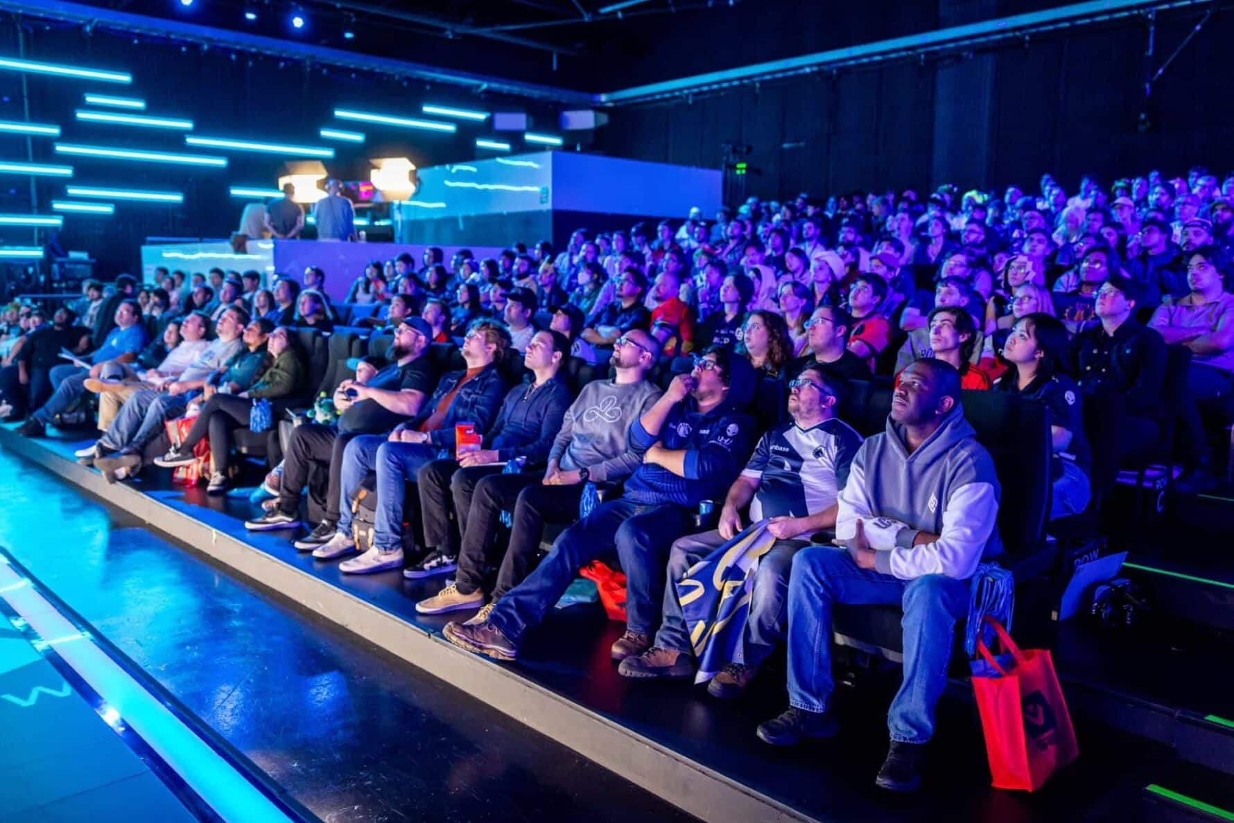 Eine Menge Zuschauer sitzt auf Sitzreihen und verfolgt aufmerksam ein Live-Event auf einer nicht sichtbaren Bühne oder Leinwand in einem dunklen, von leuchtend blauem Neonlicht beleuchteten Veranstaltungsort