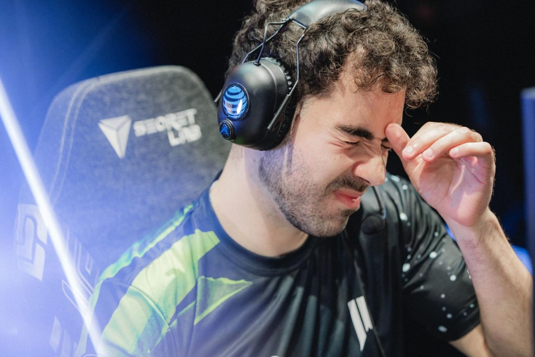 Ein dunkelhaariger, bärtiger E-Sport-Spieler sitzt in einem Secretlab-Gaming-Stuhl, trägt ein schwarz-grünes Trikot und ein Logitech G-Gaming-Headset und kneift sich verzweifelt in den Nasenrücken