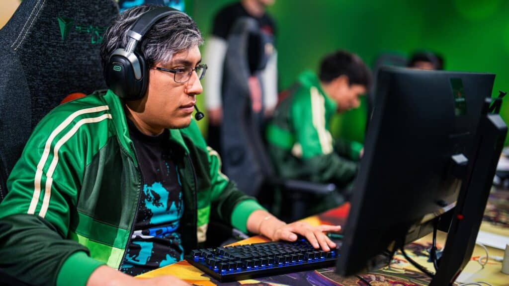 Person wearing glasses and a headset, seated at a desktop computer during an esports event, focused on gameplay while teammates sit at computers in the background.