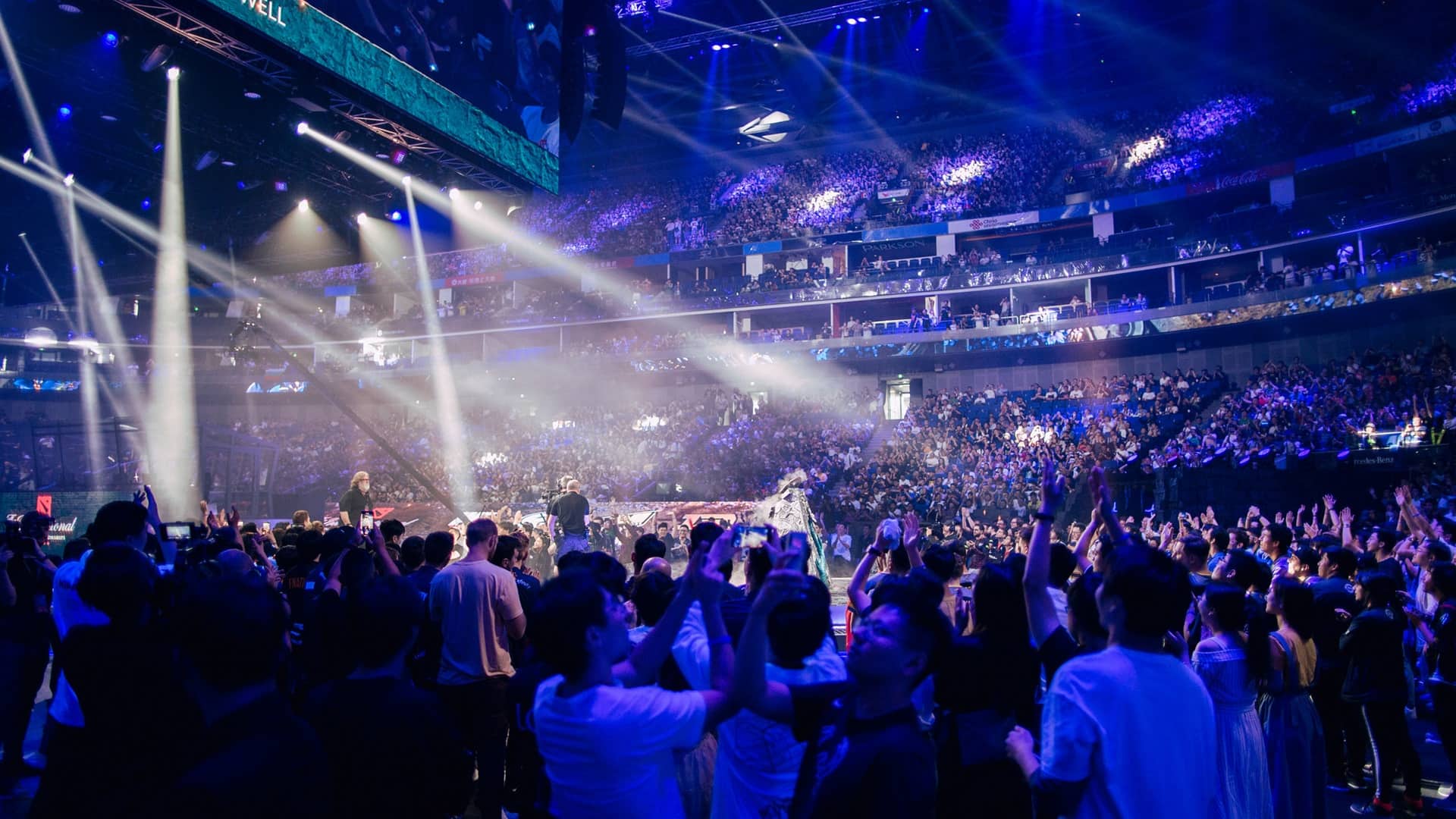 Eine weite Luftaufnahme der überfüllten Mercedes-Benz Arena in Shanghai, beleuchtet von leuchtend blauer und violetter Bühnenbeleuchtung, während der Dota 2 The International 2019 eSports-Meisterschaftsveranstaltung