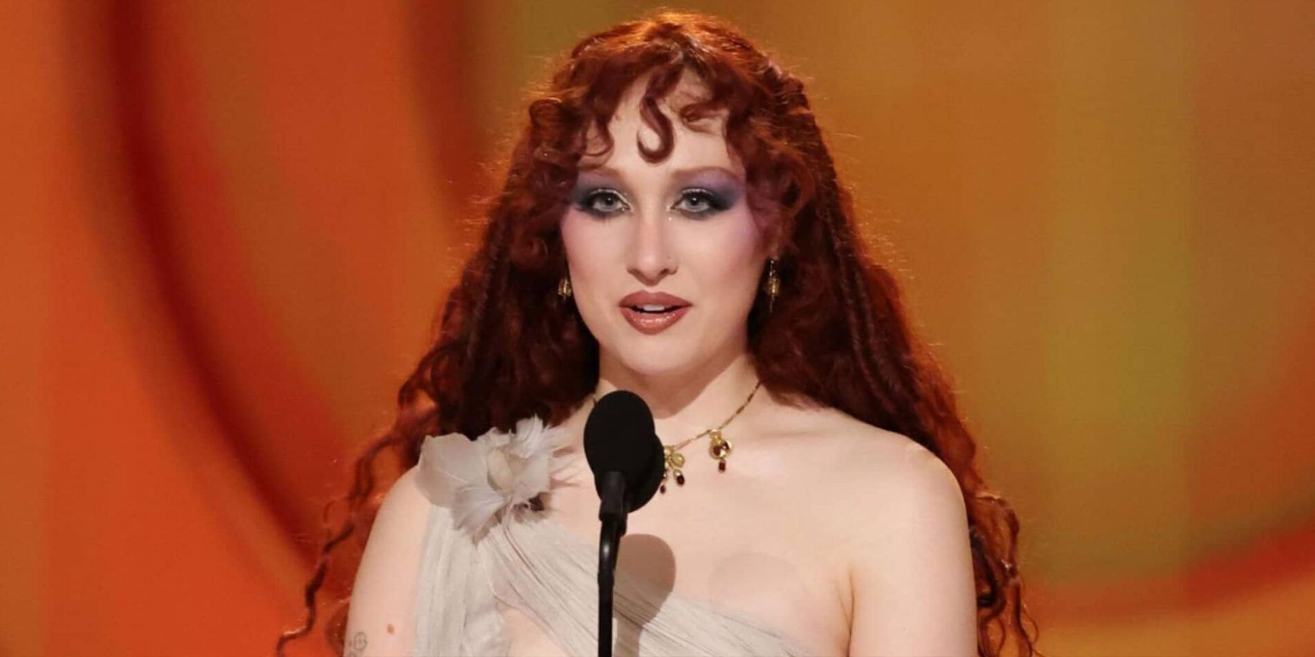 An illustration of singer Chappell Roan at an awards ceremony, featuring her distinctive long red curly hair, dramatic blue eyeshadow, and a light-colored, off-the-shoulder dress with a flower detail, as she speaks into a microphone against an orange background